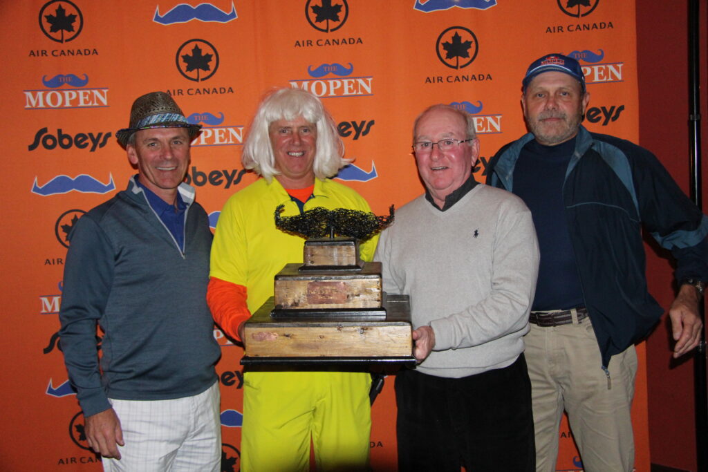 Four men posing in front of an orange MOpen backdrop, holding a large wooden trophy topped with a black mustache sculpture. One man wears a white wig and bright yellow outfit.